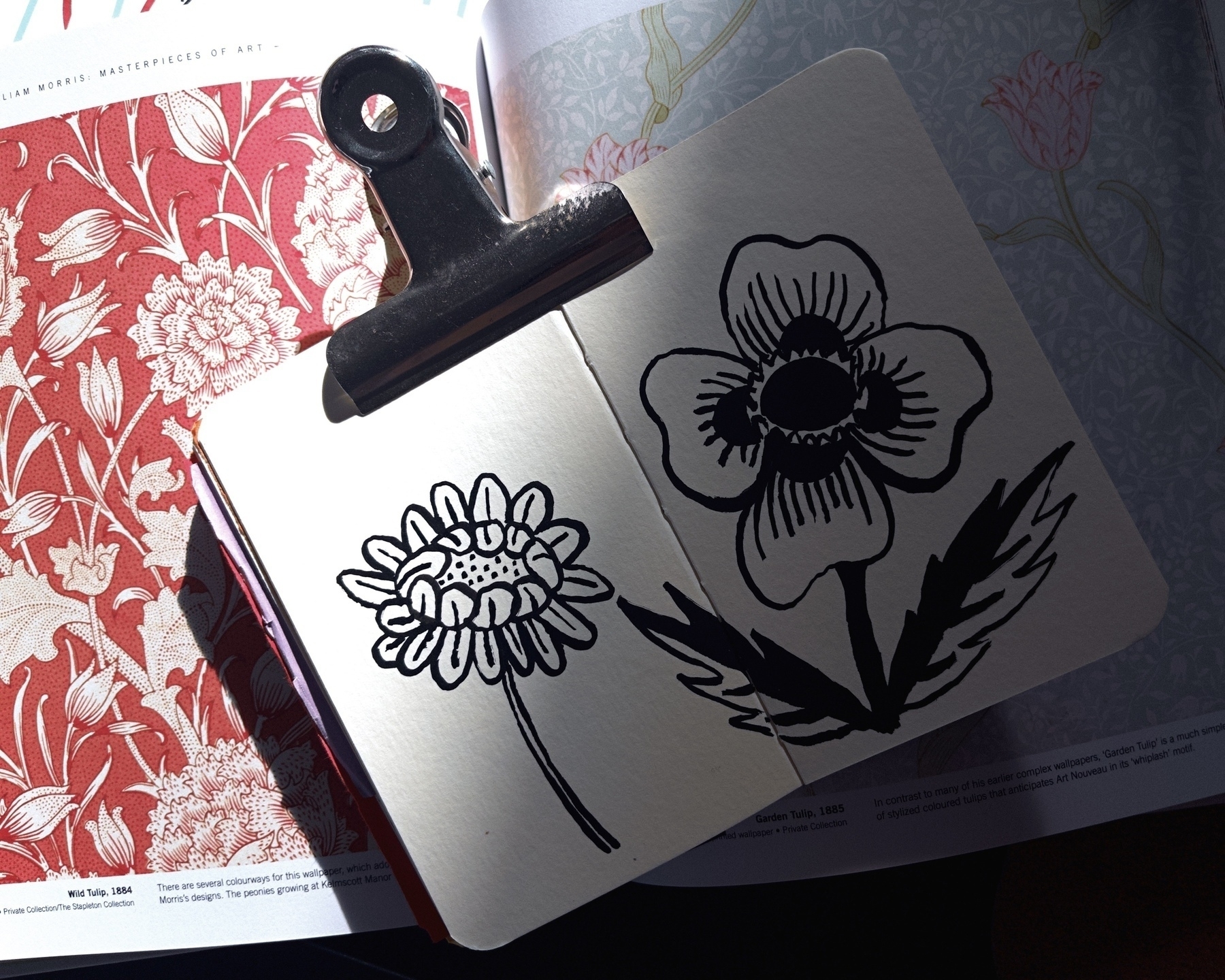 A sketchbook held open by a clip displays black ink drawings of flowers, resting on a book with floral patterns.